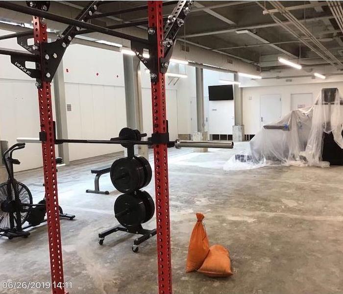 Gym with workout equipment, some wrapped in plastic, floor exposed